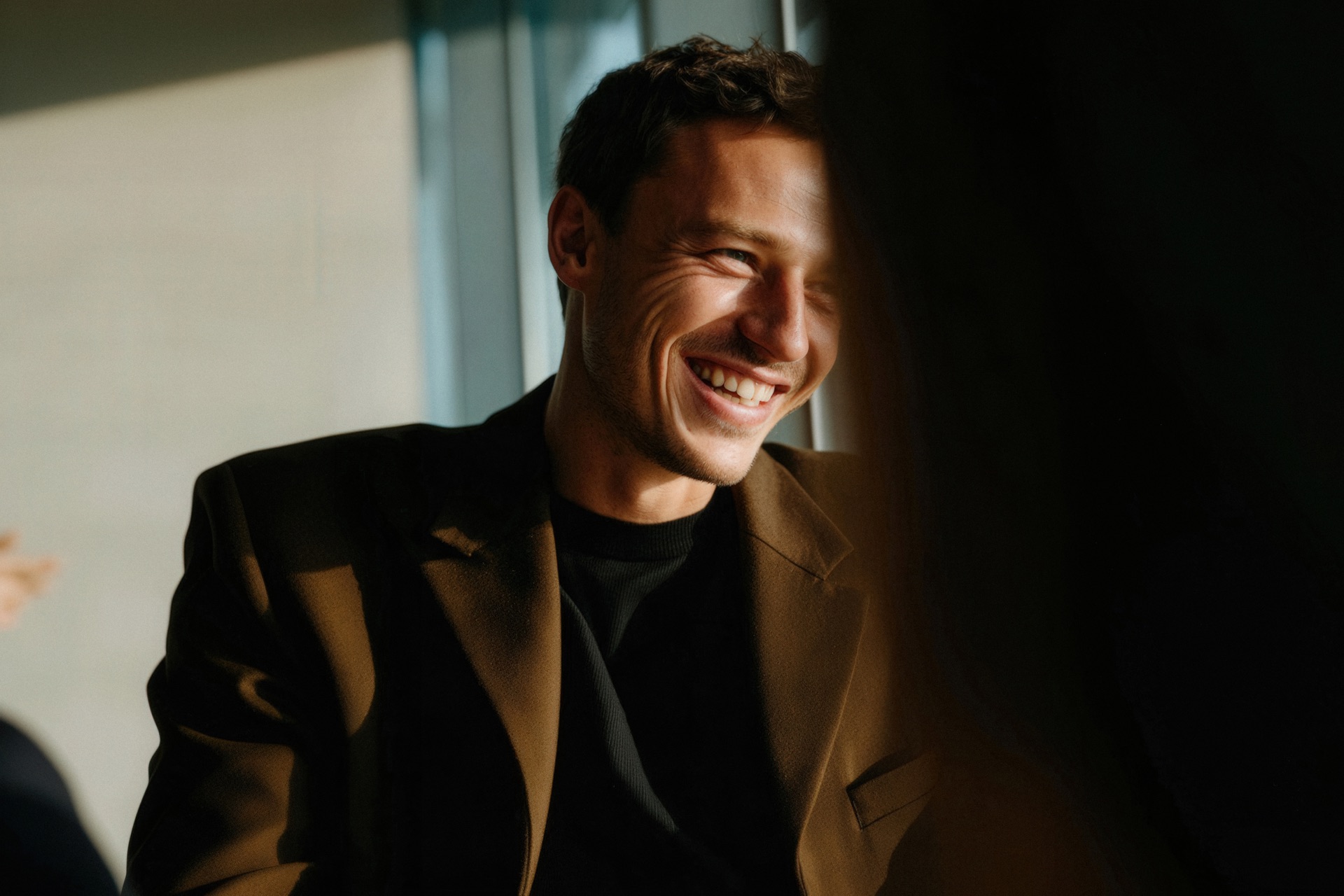 A smiling man in a brown jacket, lit by a window